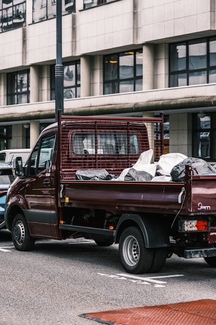 A small burgundy flatbed pickup truck parked on a city street in front of a modern office building with large glass windows and a concrete facade. The truck's cargo bed is filled with various black an